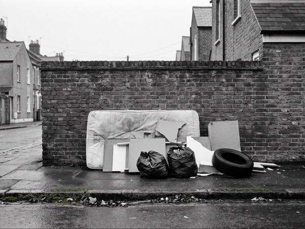 Fly-tipped mattress, furniture and bin bags dumped against a brick wall on a wet residential street